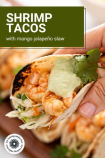 Hand holding up a shrimp taco topped with avocado crema and slaw. Text overlay includes recipe name.