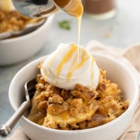 Caramel sauce being drizzled over apple pear crisp and whipped cream in a white bowl