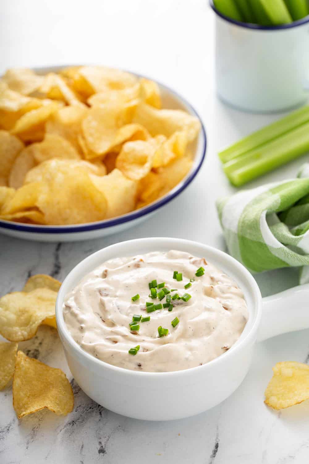 Bowl of french onion dip with potato chips and celery sticks in the background