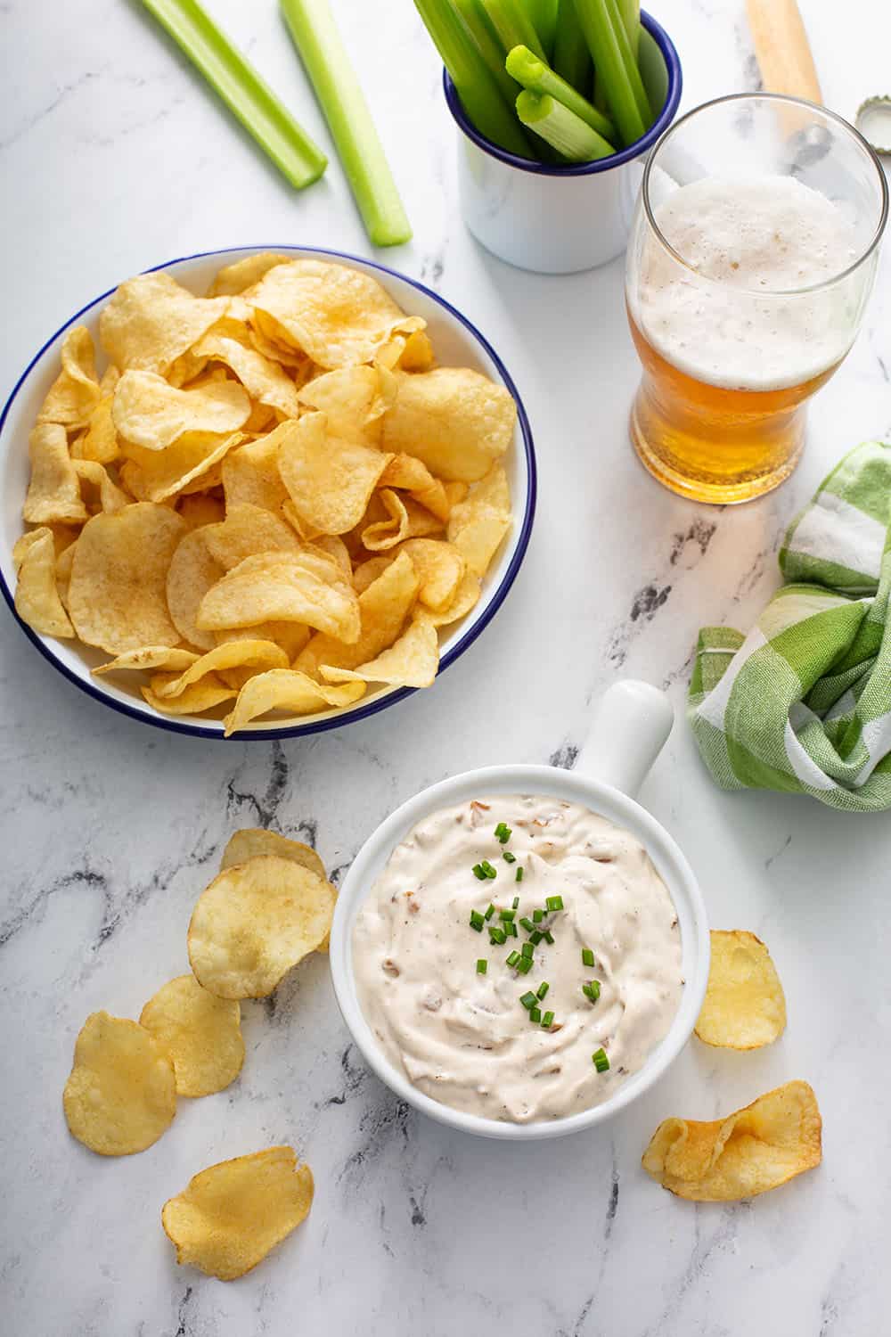 Overhead view of a bowl of french onion dip on a counter next to a bowl of potato chips and a glass of beer