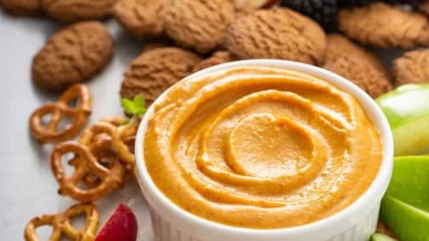 Pumpkin dip in a white bowl surrounded by fruit, pretzels, and cookies