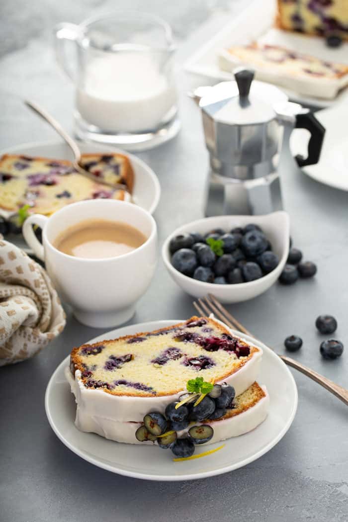Slices of lemon blueberry bread on a white plate with cups of coffee, a bowl of blueberries, and a second plate of bread in the background