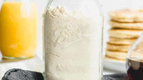 Homemade Bisquick mix in a glass jar with a stack of pancakes and a carafe of orange juice in the background