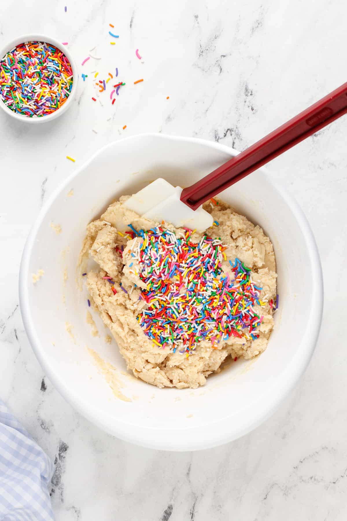 Rainbow sprinkles being added to cookie dough in a white mixing bowl.