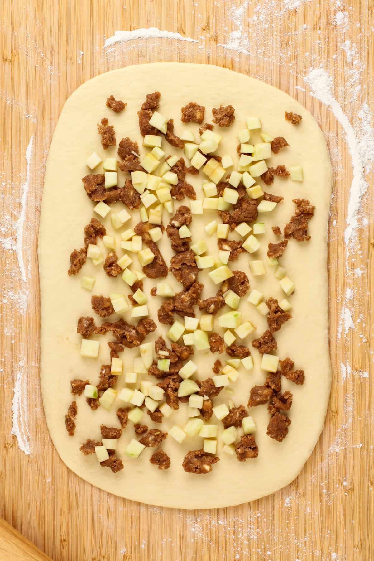 Cinnamon roll filling and apple pieces spread onto yeast dough on a wooden counter.