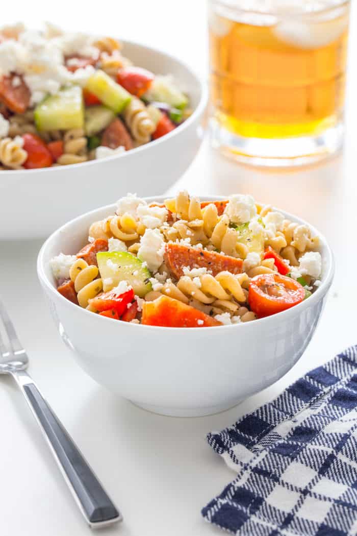 Greek pasta salad in a small white bowl next to a blue napkin on a white countertop