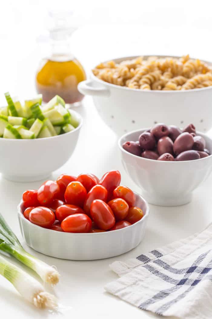 Bowls of fresh vegetables for greek pasta salad arranged on a white countertop