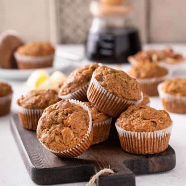 Morning glory muffins piled on a wooden board. A pot of coffee is visible in the background.