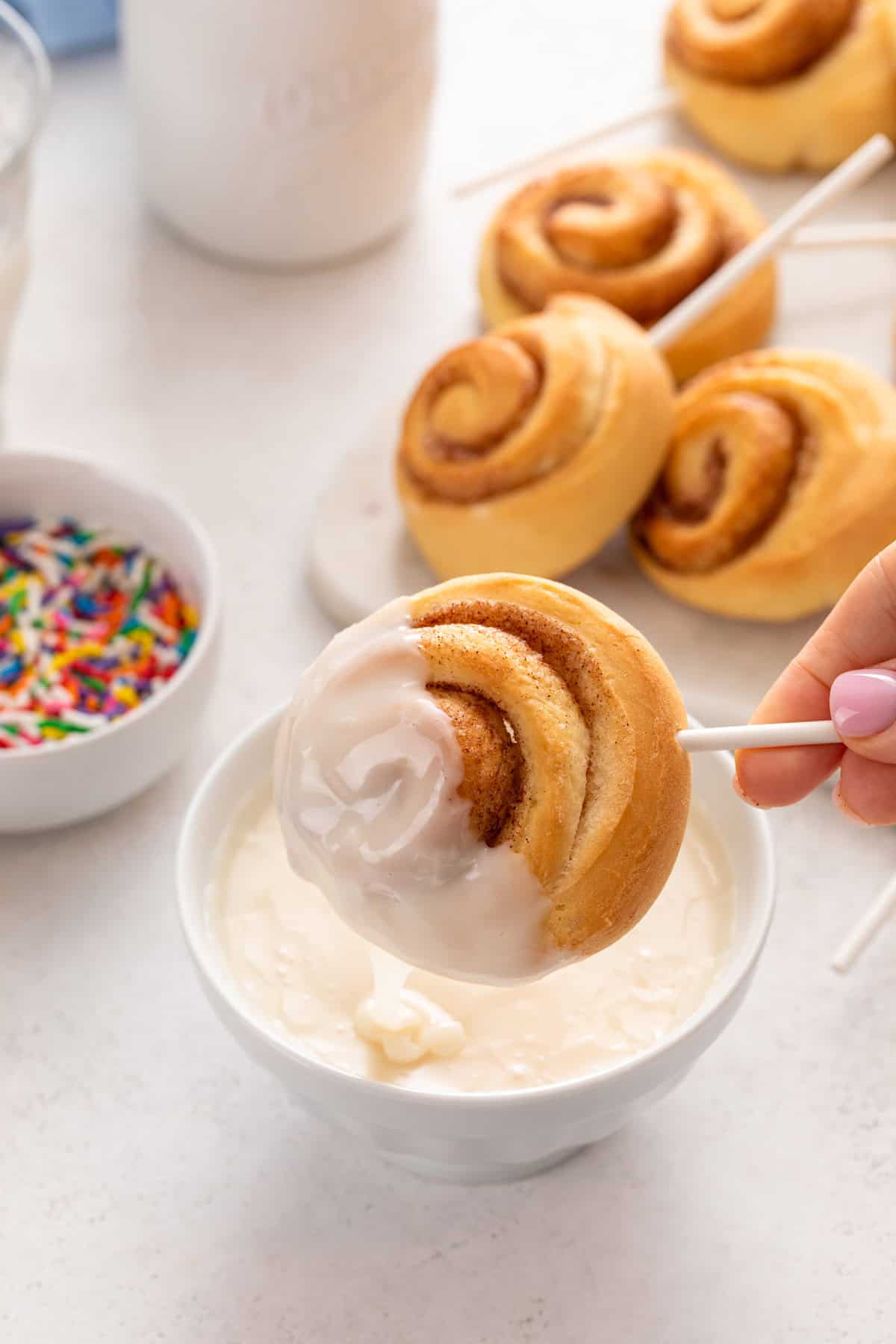 Hand dipping a mini cinnamon roll on a lollipop stick into a bowl of glaze.