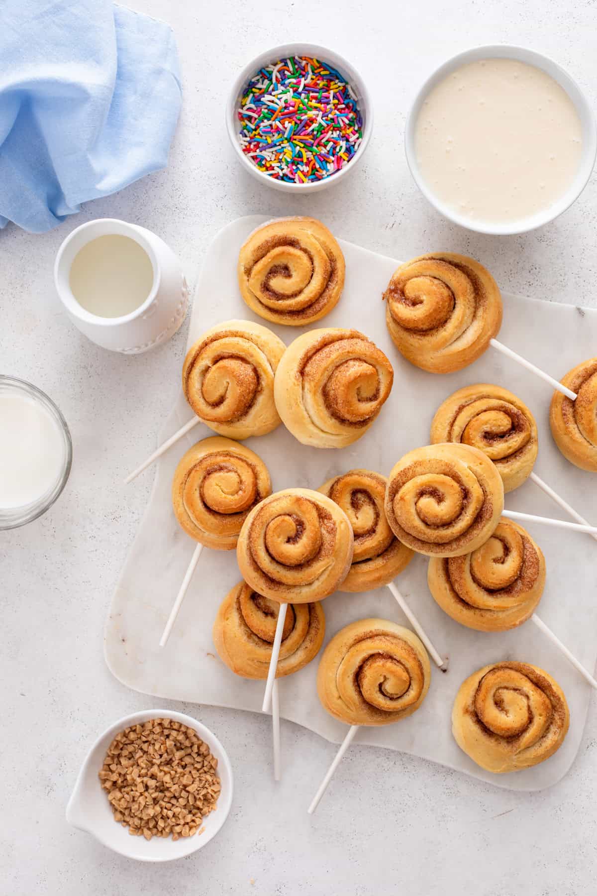 Mini cinnamon rolls on lollipop sticks with bowls of glaze and toppings around them.