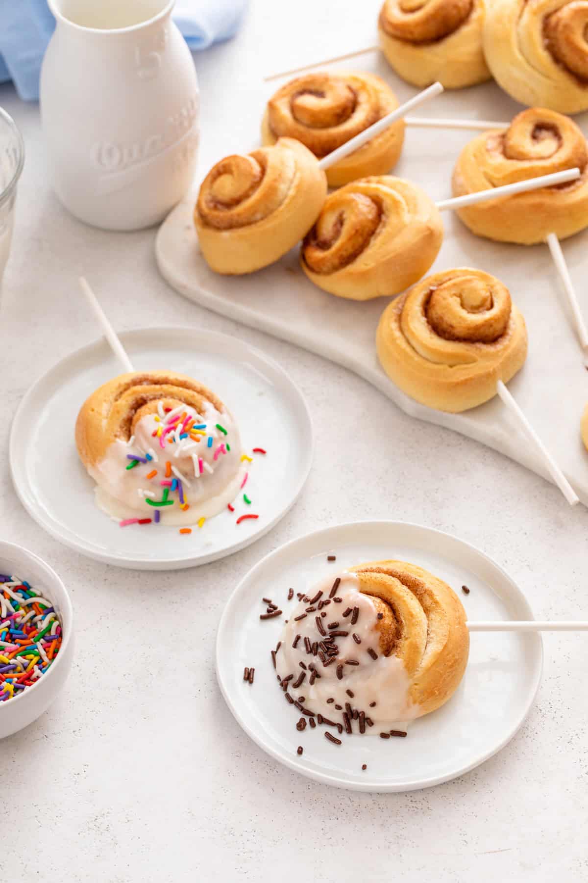 Two white plates, each holding a mini cinnamon roll on a lollipop stick dipped in glaze and topped with sprinkles.