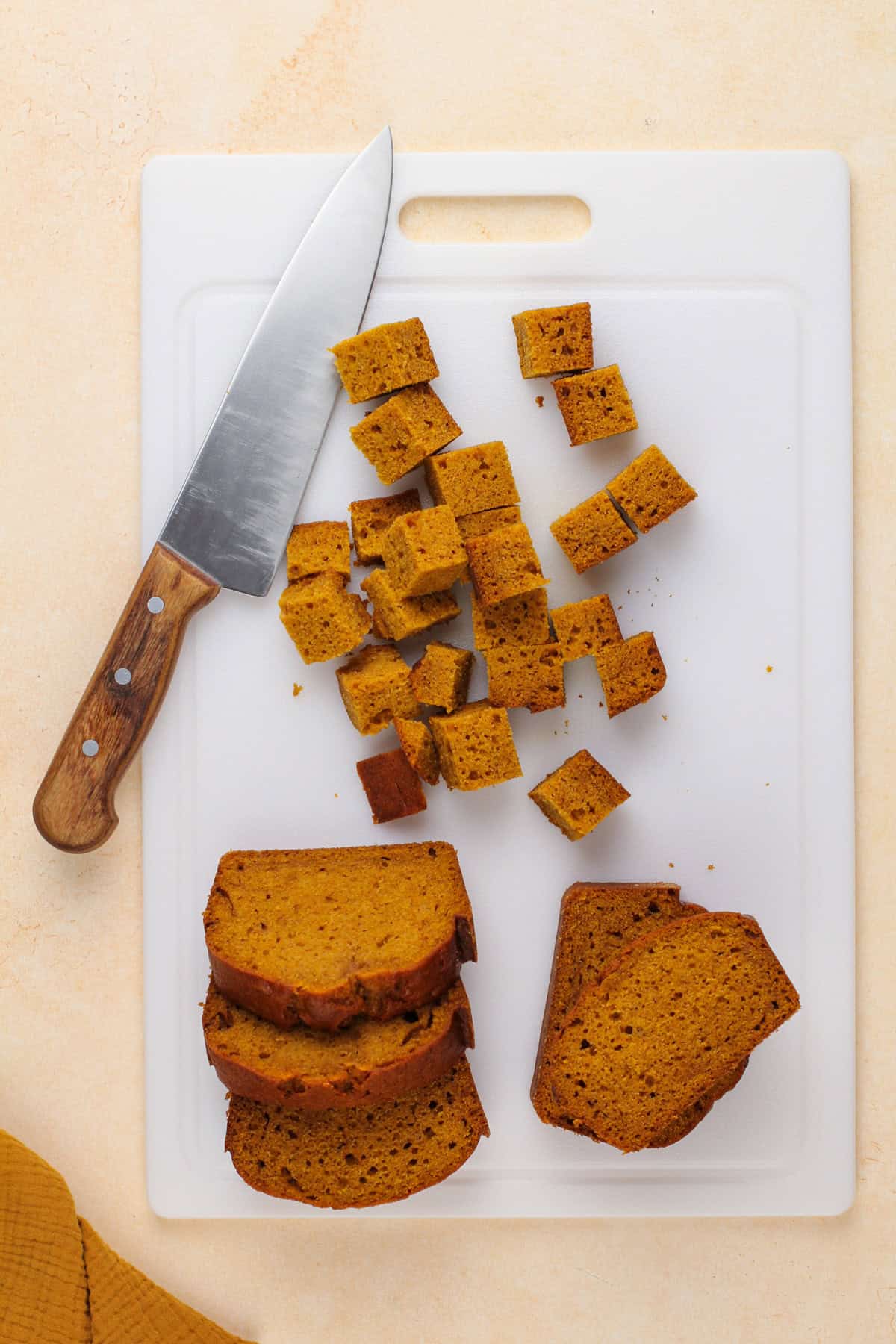 Pumpkin bread being cut into cubes on a white cutting board.