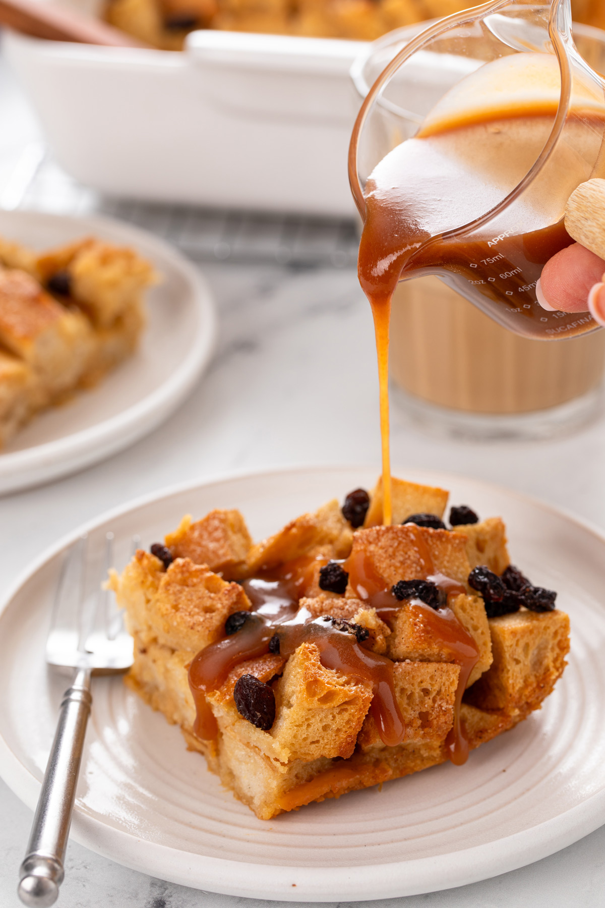 Whiskey caramel sauce being poured from a pitcher over a slice of irish bread pudding on a white plate.