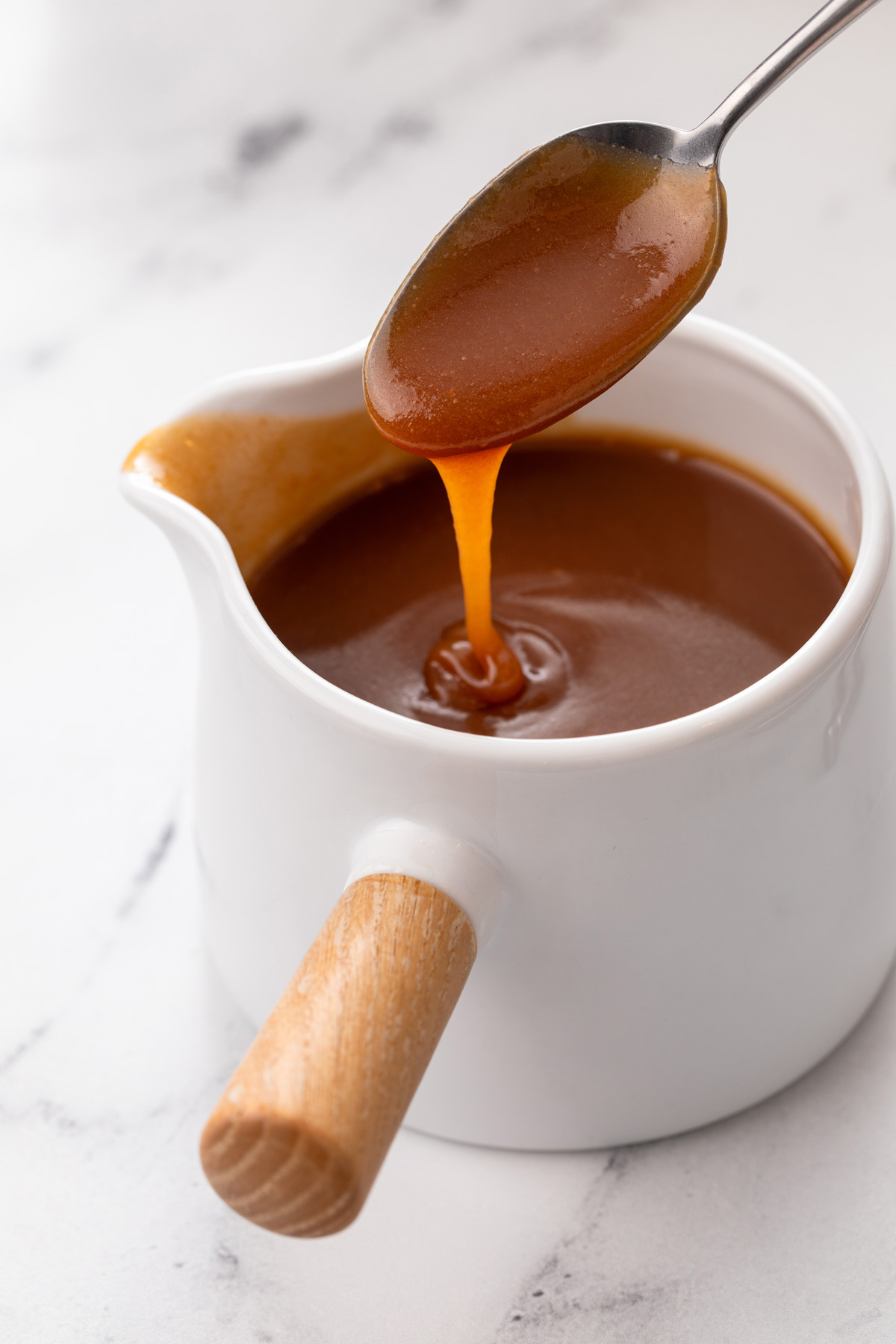 Spoon drizzling whiskey caramel sauce into a white pitcher of the sauce.