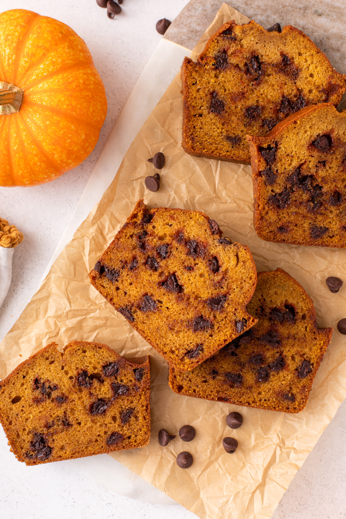 Several slices of chocolate chip pumpkin bread scattered on a piece of parchment paper next to a pumpkin.