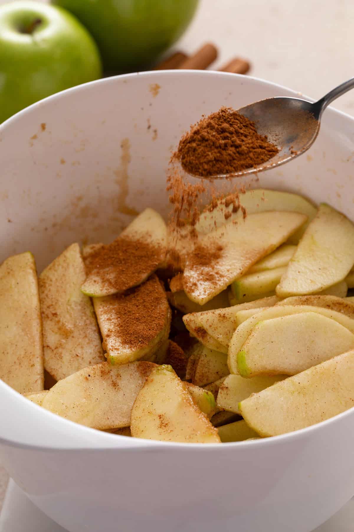 Spoon sprinkling apple pie spice over sliced apples in a bowl.