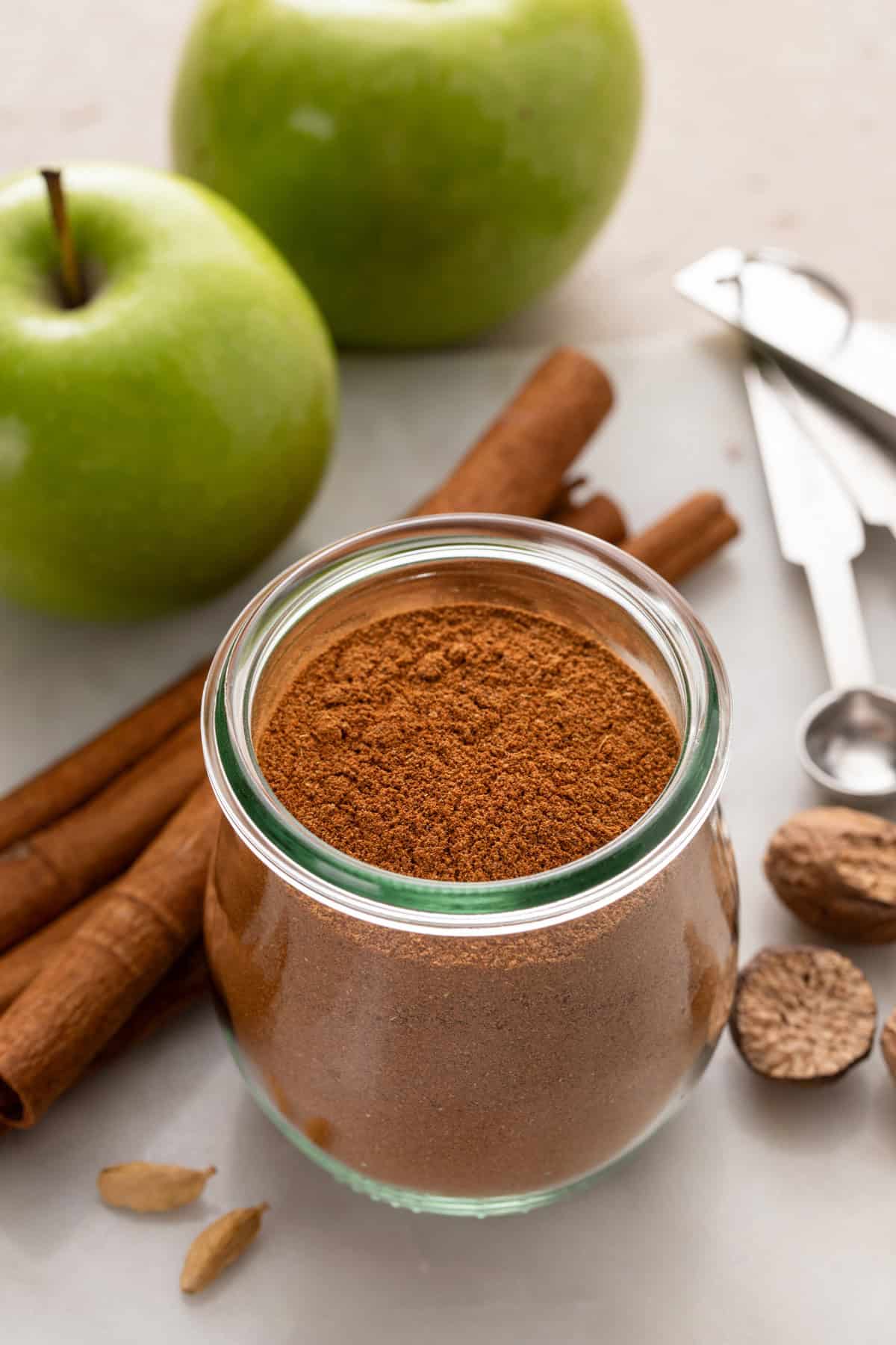 Jar filled with apple pie spice surrounded by cinnamon sticks, nutmeg, and apples.