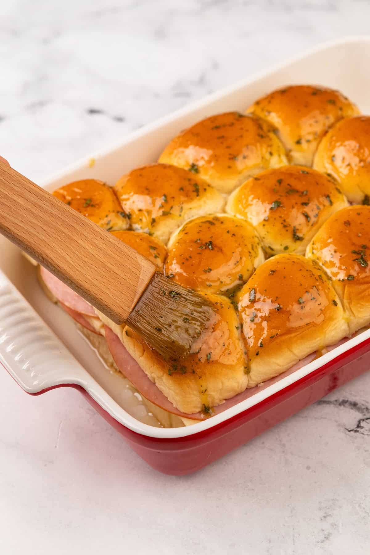 Herb butter being brushed on top of ham and cheese sliders before baking.