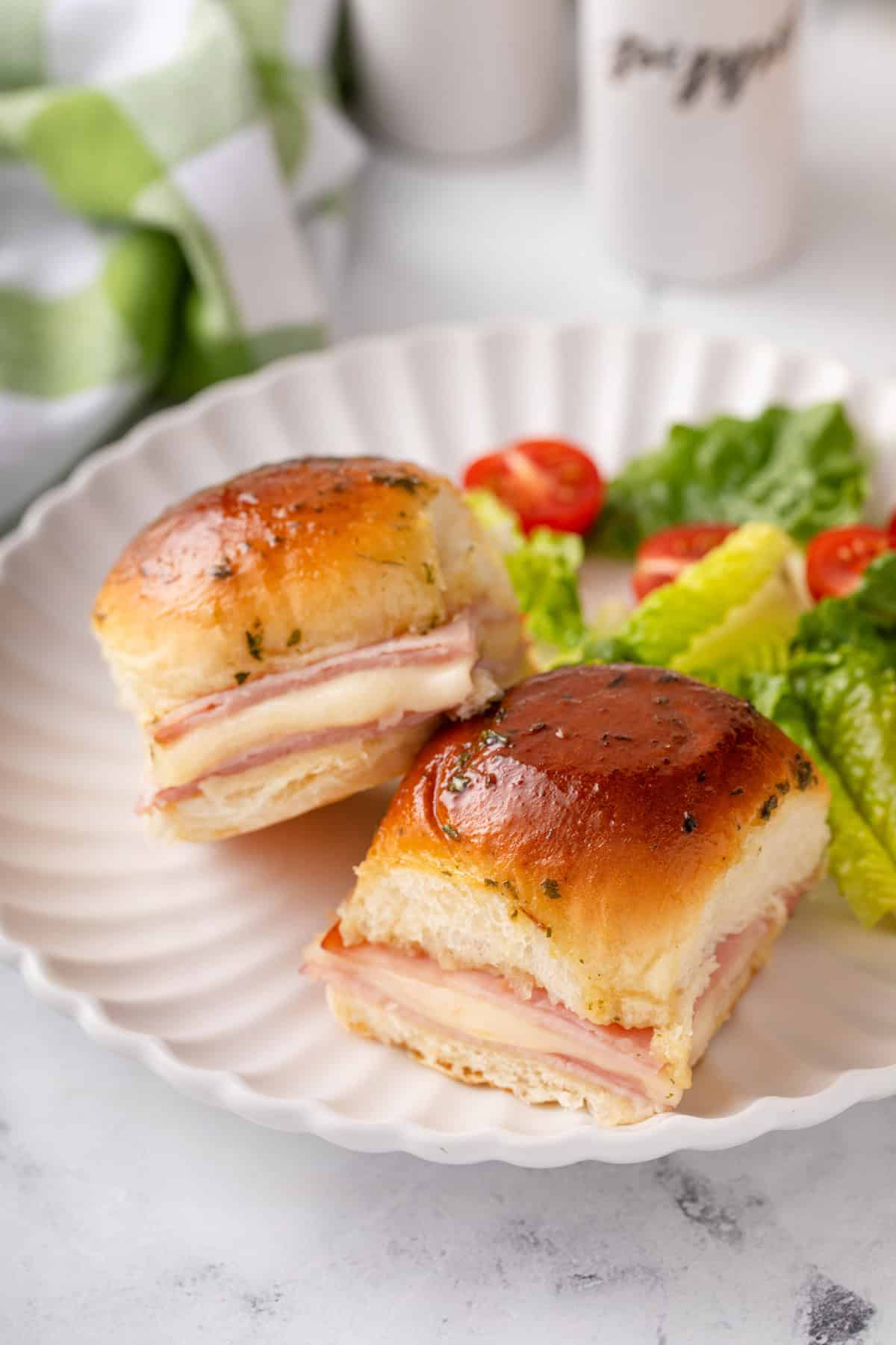 Close up of two hot ham and cheese sliders and a side salad on a white plate.