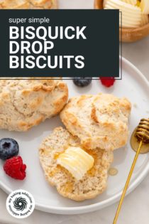 Two bisquick biscuits on a white plate next to fresh fruit. One of the biscuits is halved with butter and honey on it. Text overlay includes recipe name.