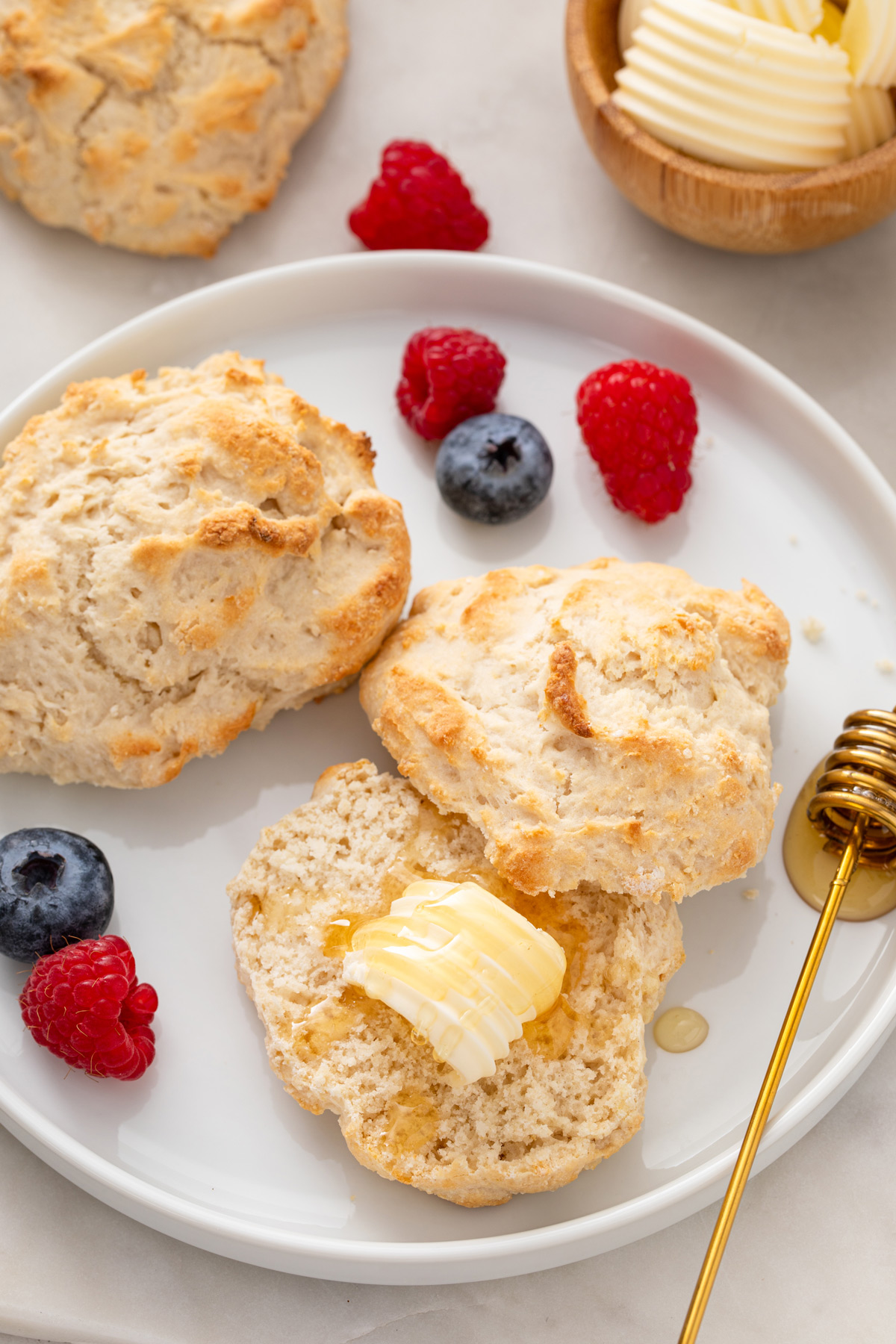 Two bisquick biscuits on a white plate next to fresh fruit. One of the biscuits is halved with butter and honey on it.