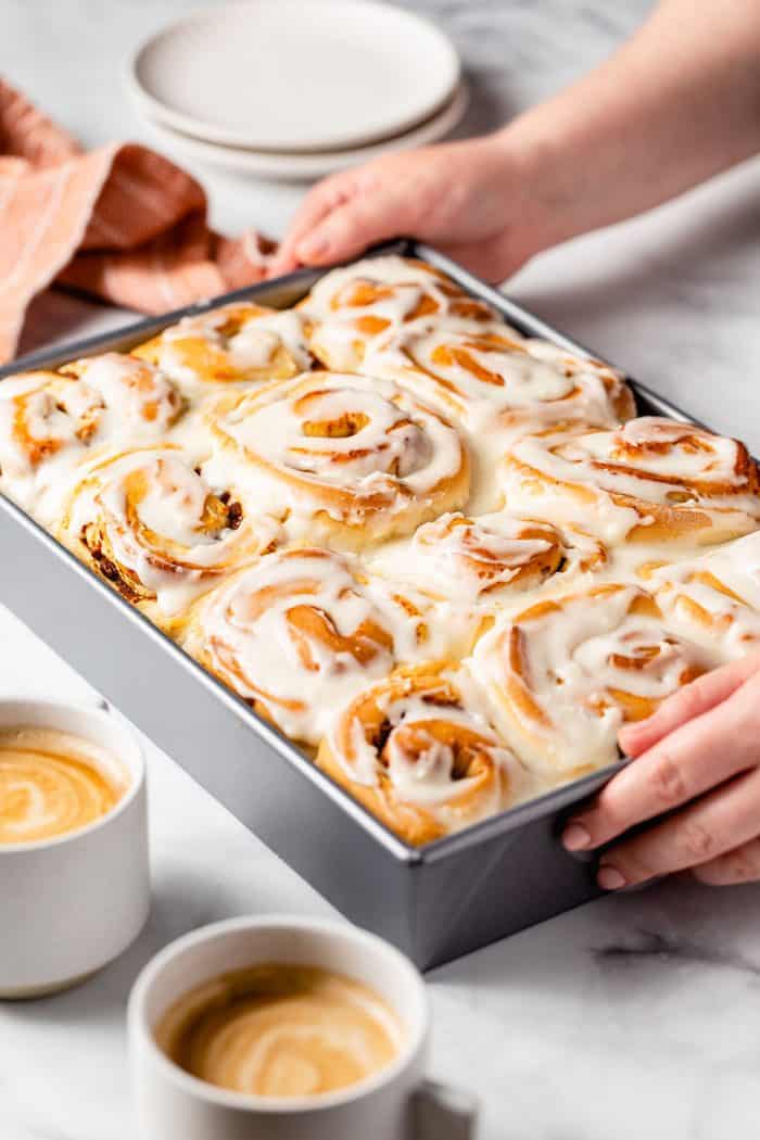 Hands placing a pan of frosted pumpkin spice cinnamon rolls on a marble countertop