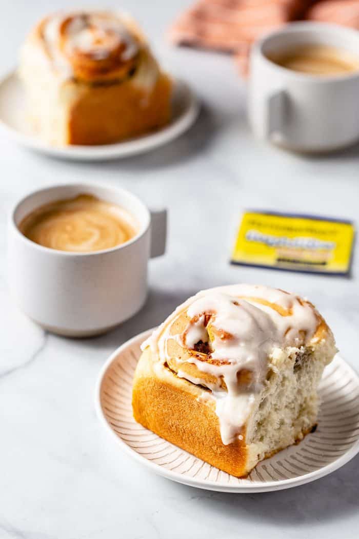 Frosted pumpkin spice cinnamon roll on a white plate with a cup of coffee in the background