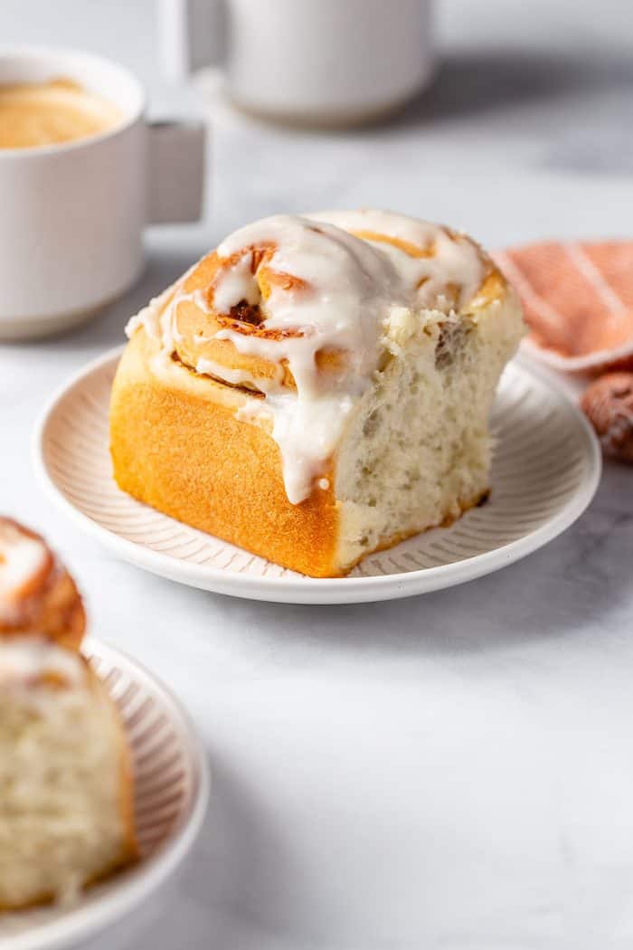 Frosted pumpkin spice cinnamon roll on a white plate with a coffee cup in the background