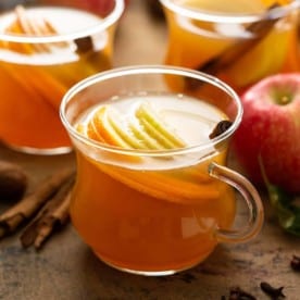 Mug of wassail with apples, cinnamon sticks, and two more mugs of wassail in the background