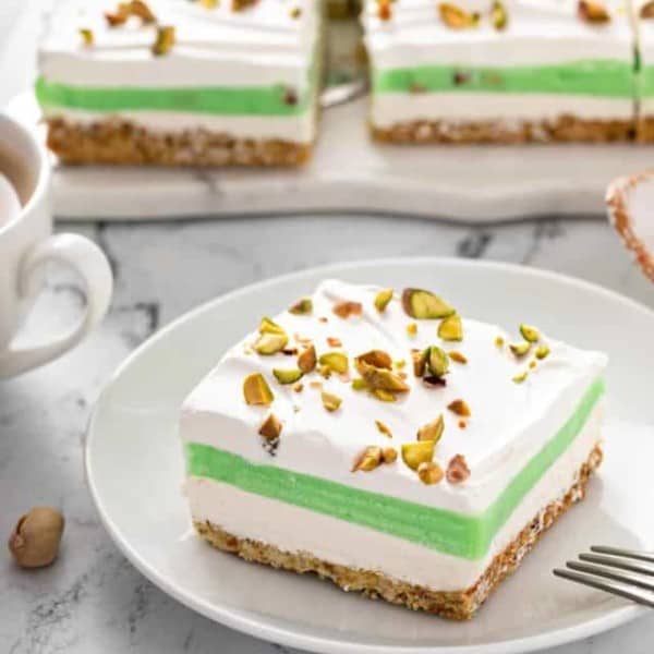 Slice of pistachio pudding dessert on a white plate, with the rest of the layered dessert in the background.