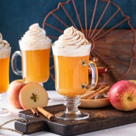 Glasses of spiced apple cider topped with whipped cream and surrounded by cinnamon sticks.