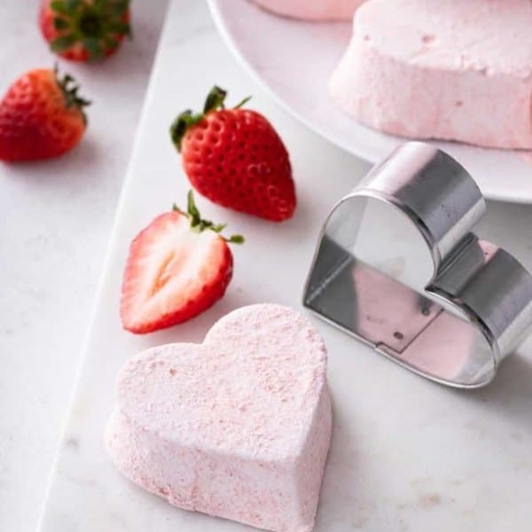 Heart-shaped strawberry marshmallow next to a metal heart cookie cutter on a white board.