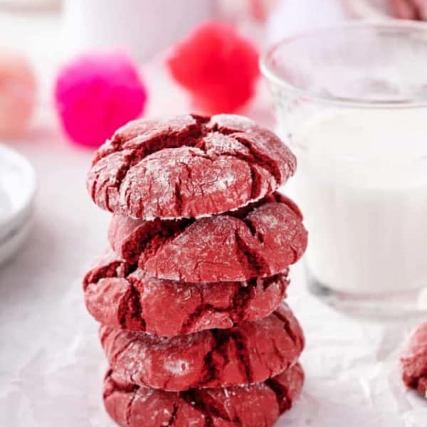 Five red velvet crinkle cookies stacked on a piece of parchment in front of a glass of milk.