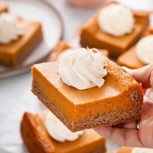 Hand holding up a slice of pumpkin gooey butter cake topped with a dollop of whipped cream.