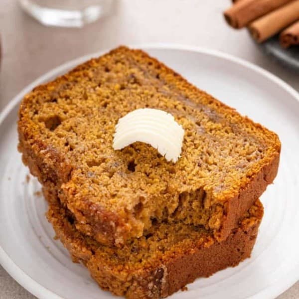 Two slices of pumpkin banana bread on a white plate. The top slice has a pat of butter and a bite taken from the corner.