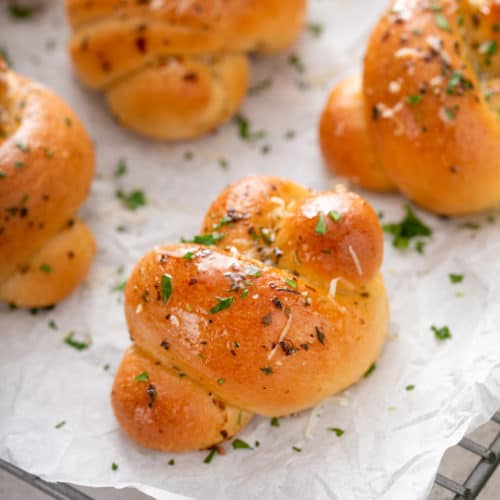 Close up of garlic knots on a piece of parchment paper, topped with parmesan cheese and fresh parsley.