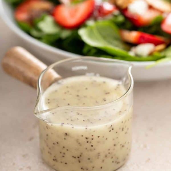 Close up of a small jar of poppy seed dressing with a bowl of strawberry spinach salad in the background.