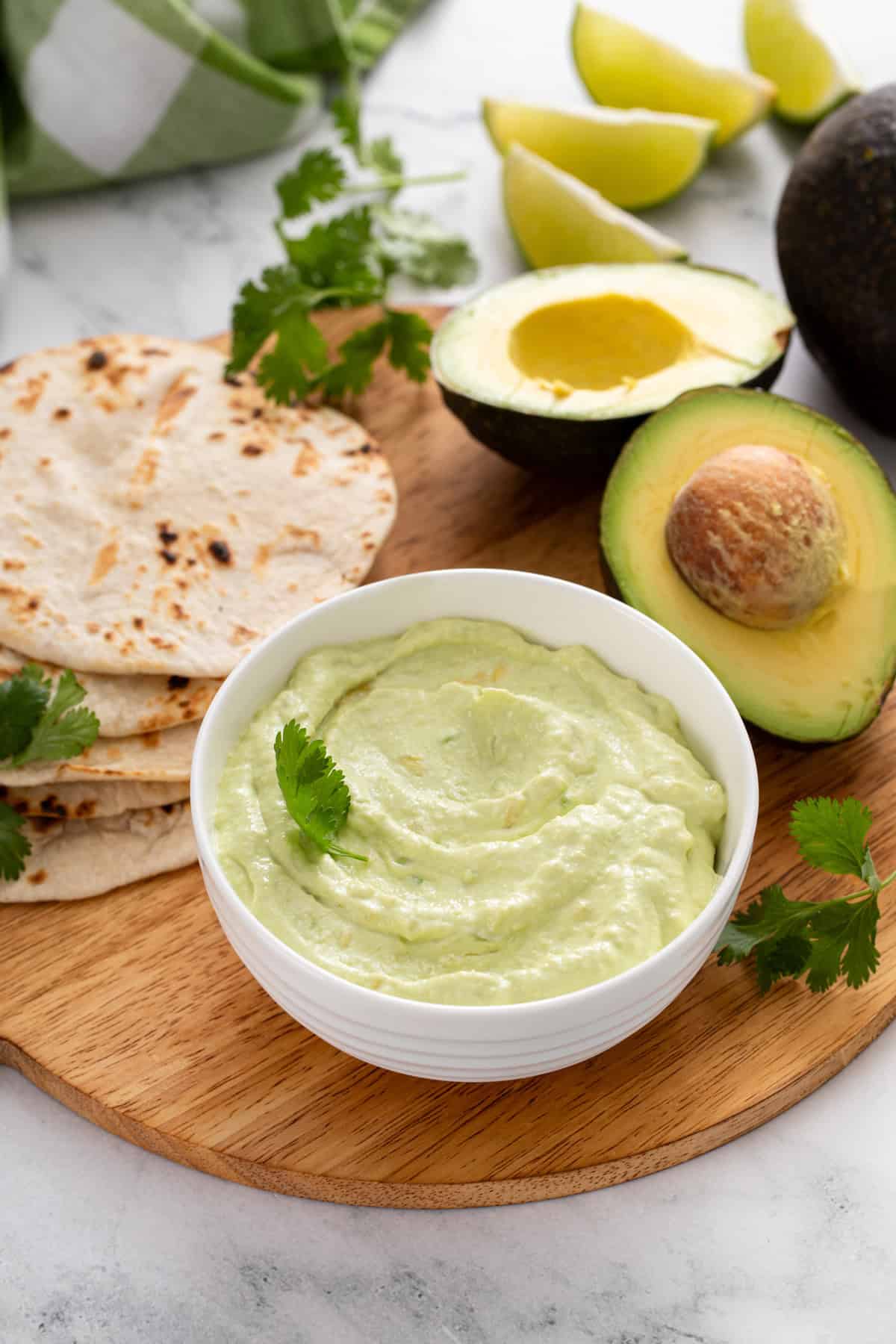 Bowl of avocado crema on a wooden platter with toasted tortillas and a halved avocado.