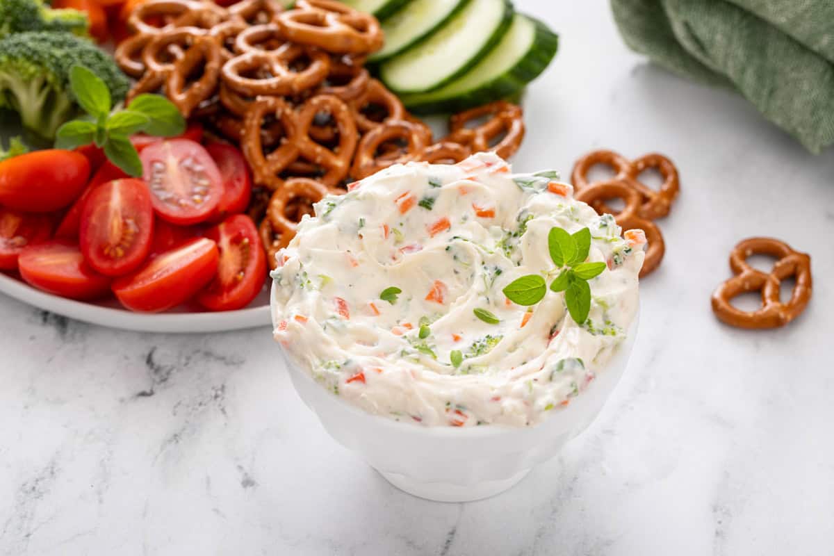 Veggie cream cheese in a white bowl with a platter of crudites and pretzels in the background.