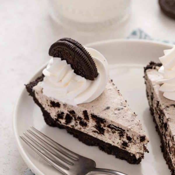 Close up of a slice of oreo pie topped with whipped cream and an oreo cookie on a white plate.