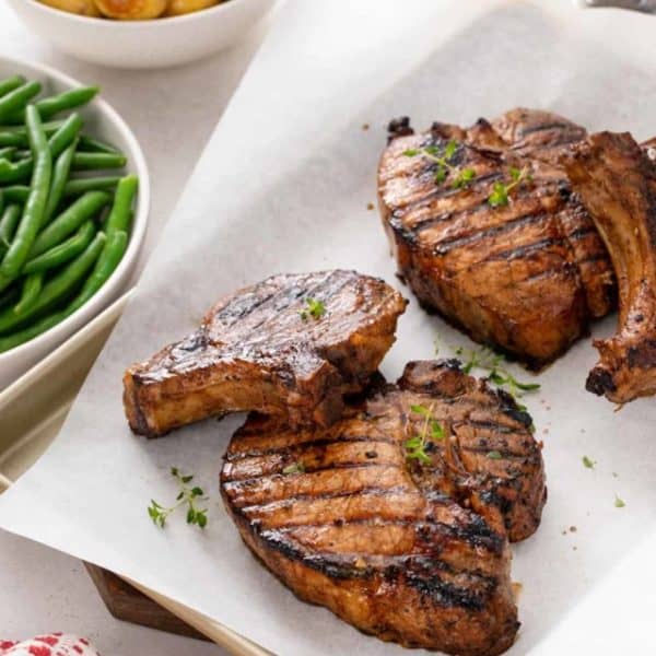 Sheet pan holding 4 grilled pork chops next to bowls of green beans and roasted potatoes.