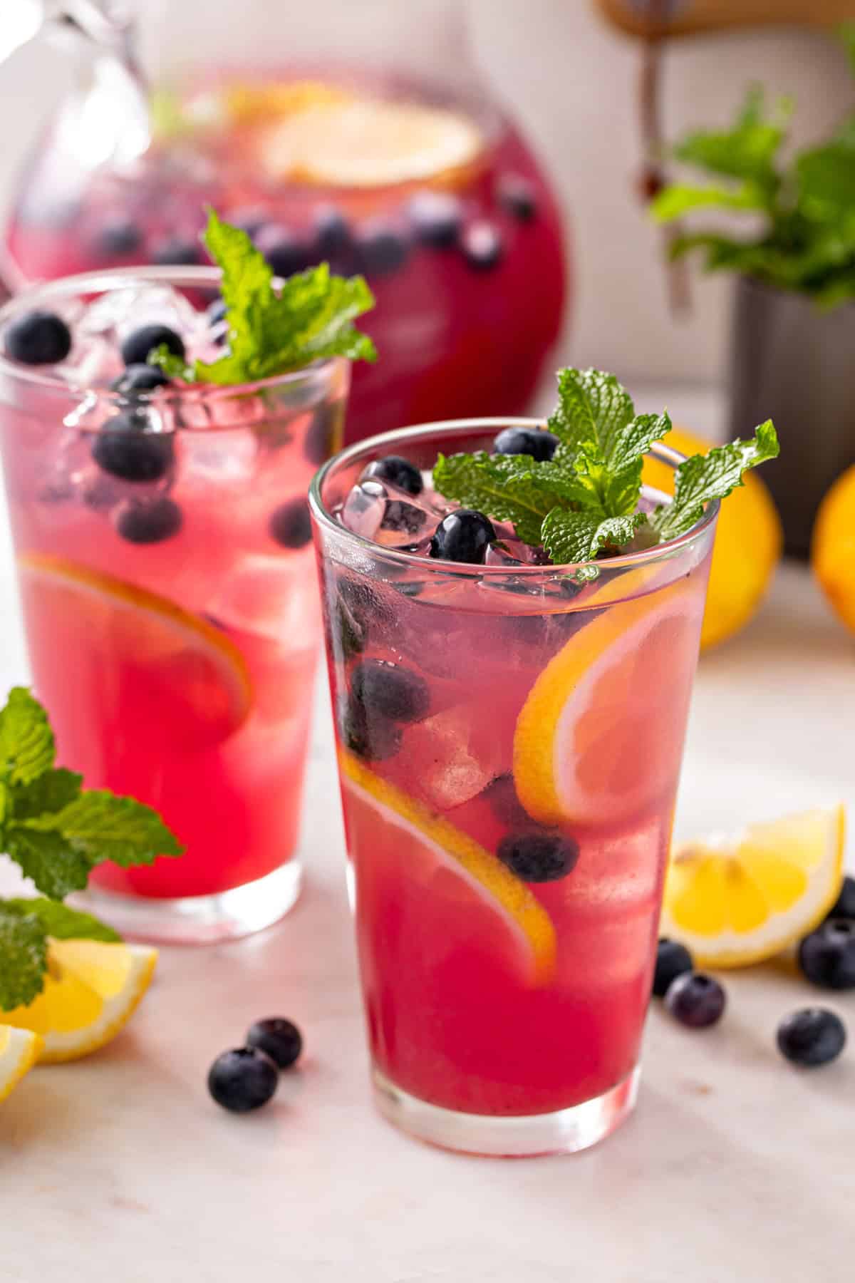 Two glasses of blueberry lemonade on a countertop with lemon wedges and fresh blueberries.