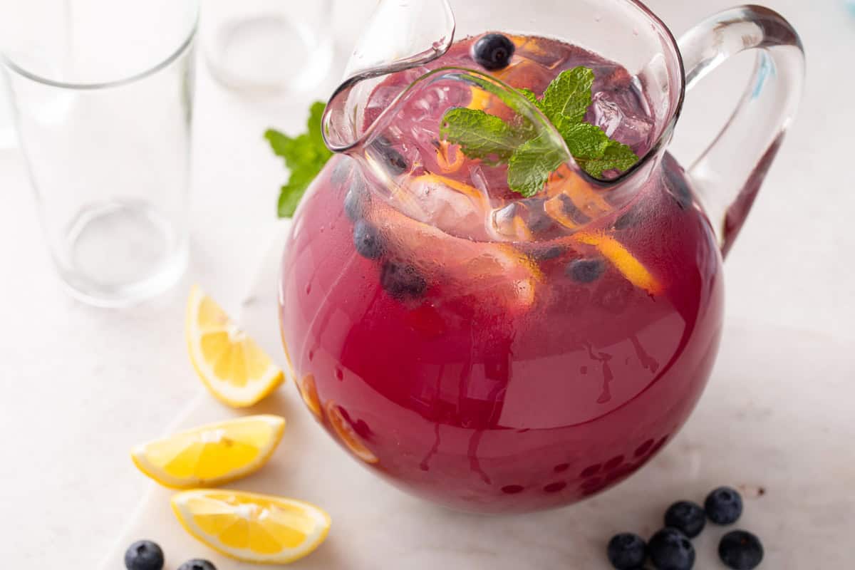 Pitcher of blueberry lemonade on a countertop next to lemon wedges.