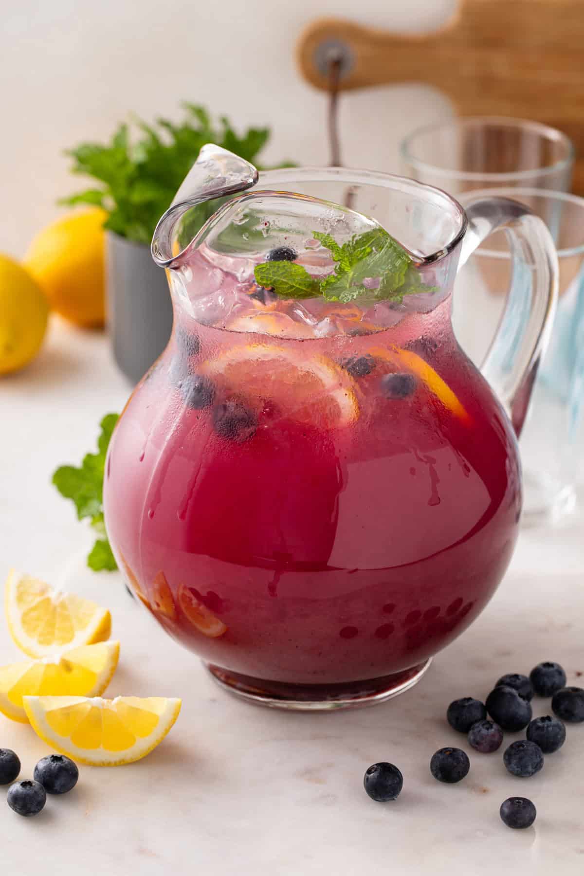 Glass pitcher filled with blueberry lemonade and garnished with lemon slices, fresh blueberries, and mint.