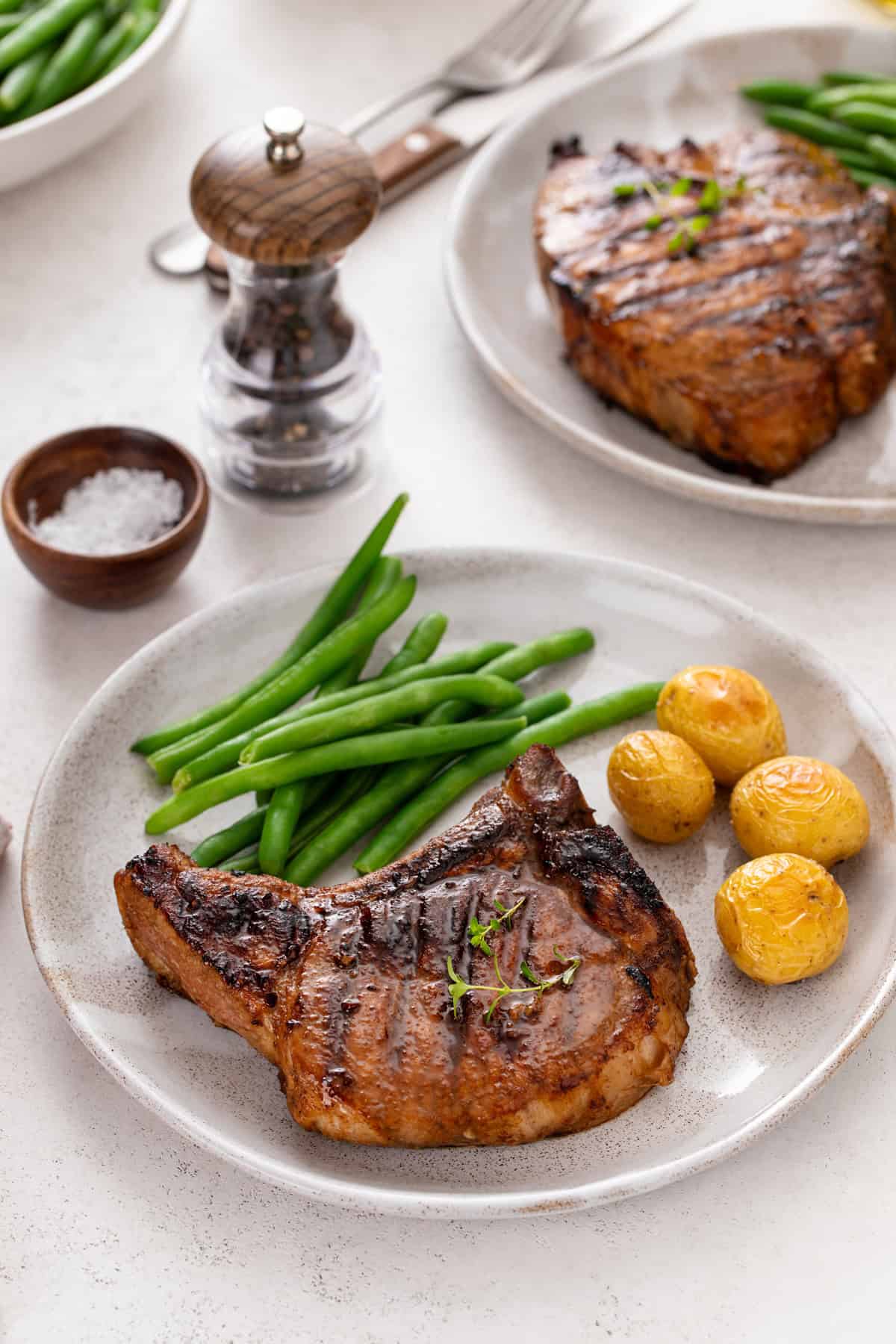 Grilled pork chop on a plate next to roasted potatoes and green beans. A second plate is visible in the background.