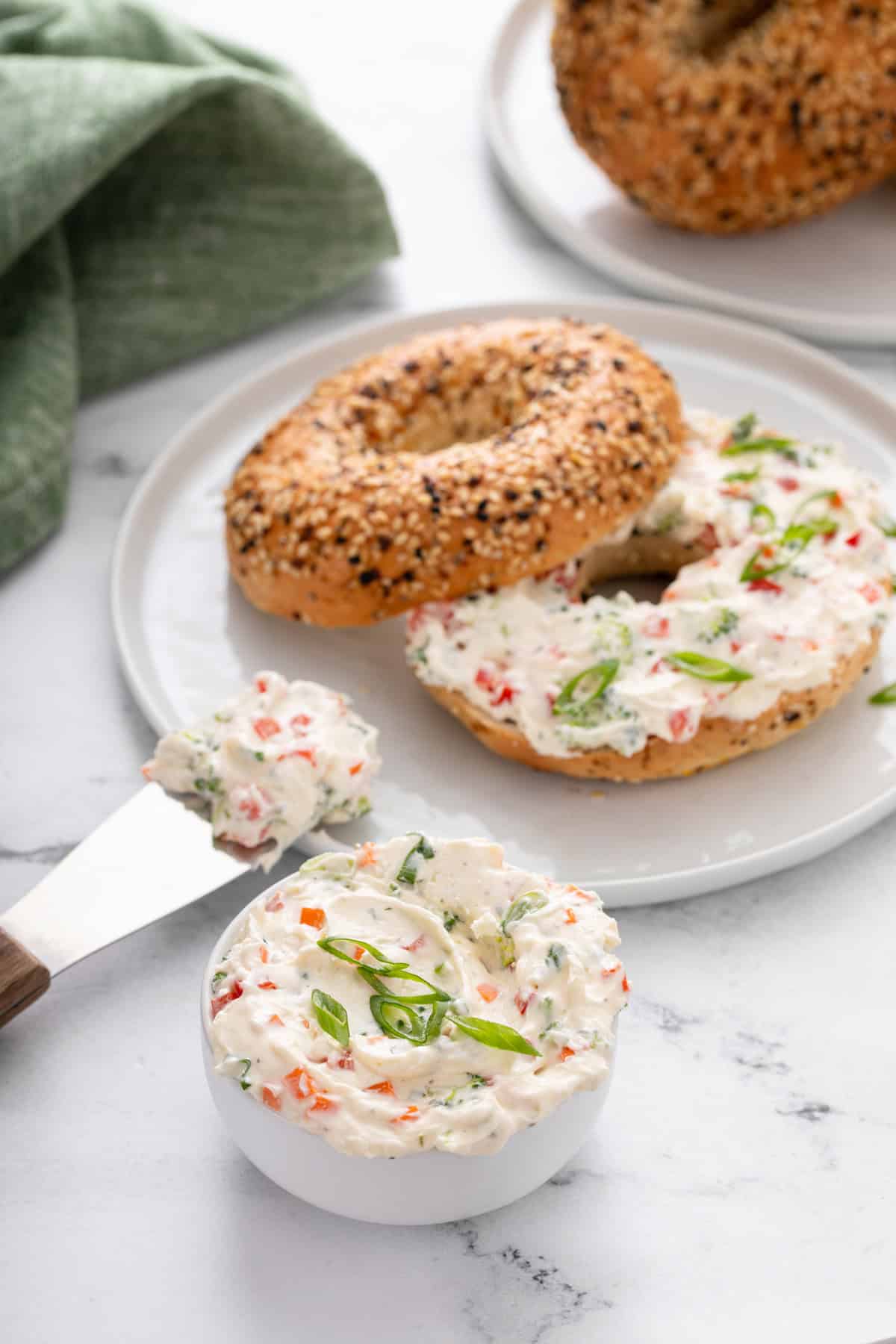 Bowl of veggie cream cheese in front of a plate with a bagel and veggie cream cheese on it.