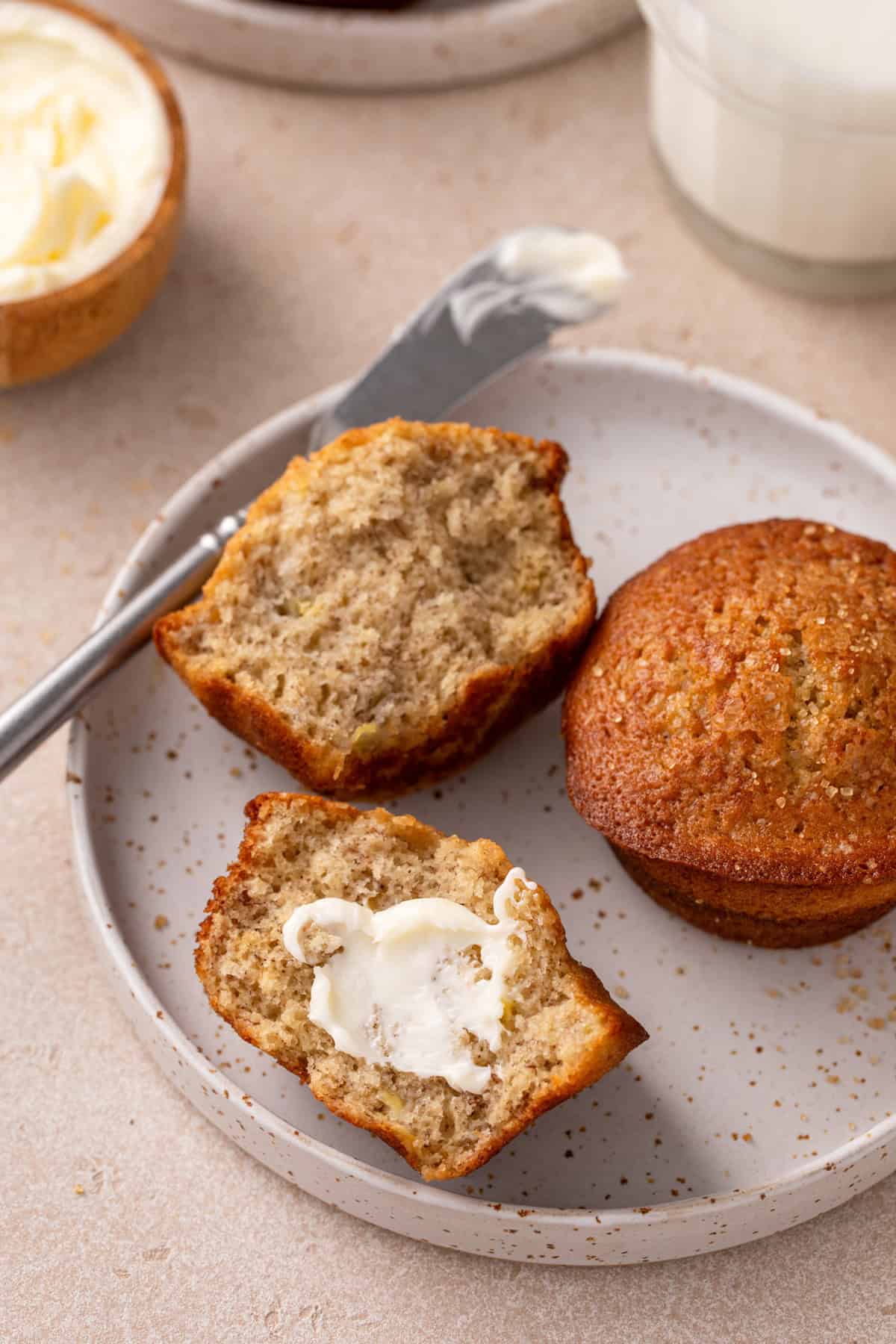 Halved and buttered banana bread muffin on a plate next to a whole banana bread muffin.