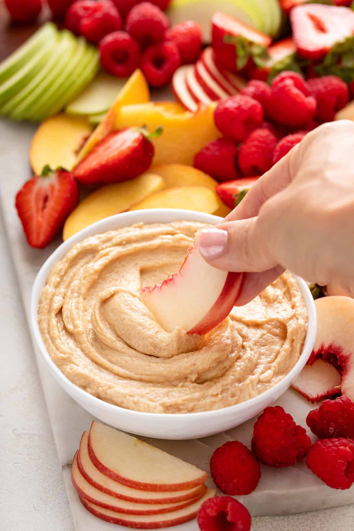 Hand dipping a slice of white peach into a bowl of peanut butter dip.