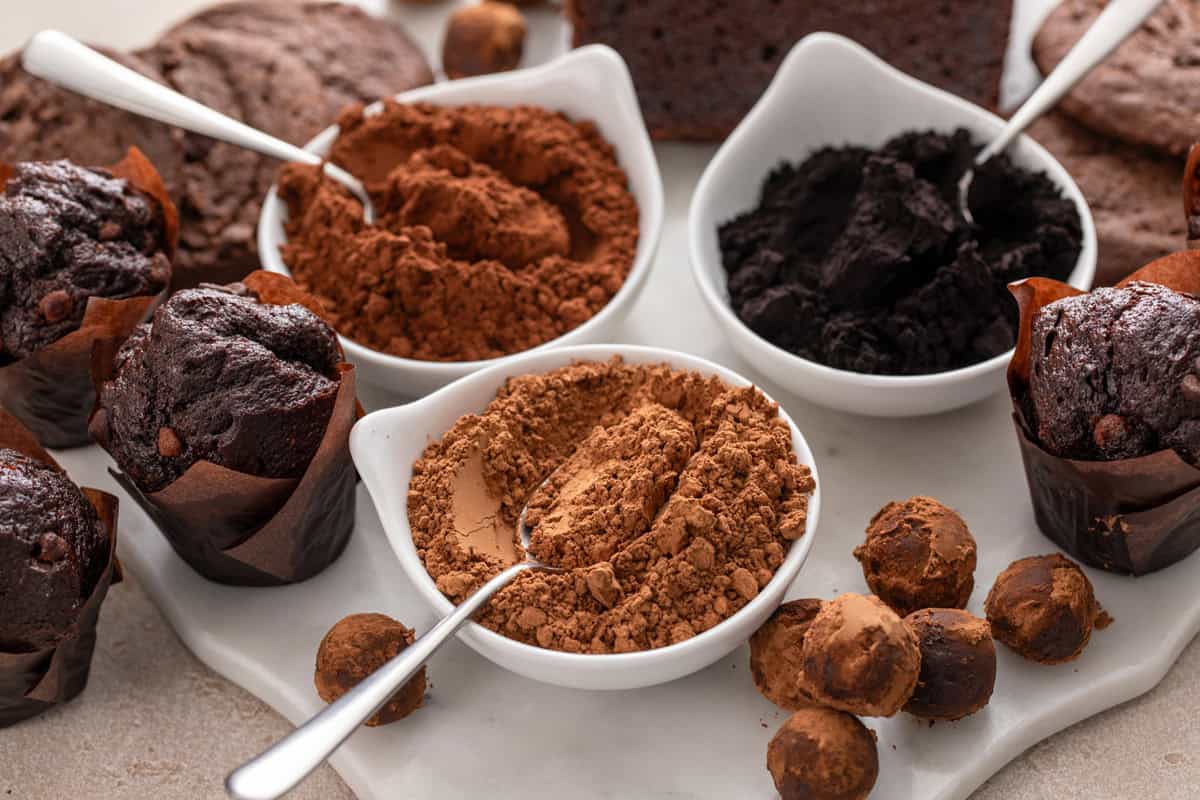 Three white bowls next to each other, each one holding natural cocoa, dutched cocoa, or black cocoa powder. The bowls are surrounded by chocolate baked goods.