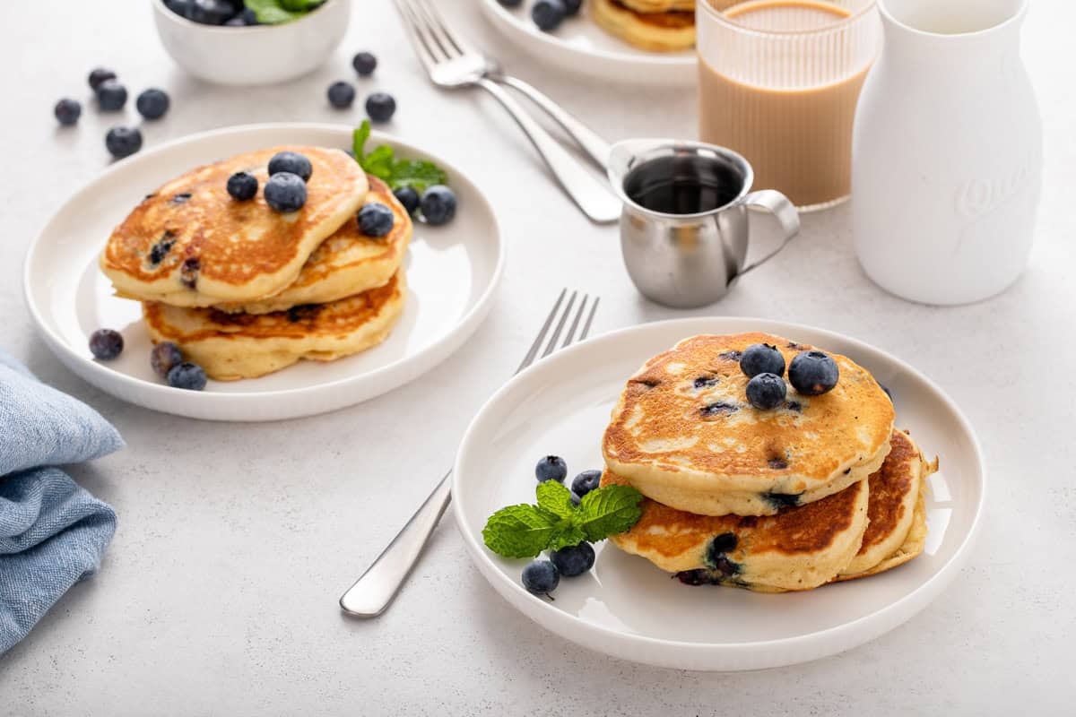 Two white plates, each holding a small stack of blueberry pancakes.